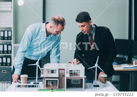 Engineers meeting about wind turbine model and house model. Renewable energy and housing development concept. Team of architects and engineers in modern office discussing design structure. Engineers meeting about wind turbine model and house model. Renewable energy and housing development concept. Team of architects and engineers in modern office discussing design structure. 119733222