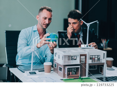 Engineers meeting about wind turbine model and house model. Renewable energy and housing development concept. Team of architects and engineers in modern office discussing design structure. Engineers meeting about wind turbine model and house model. Renewable energy and housing development concept. Team of architects and engineers in modern office discussing design structure. 119733225