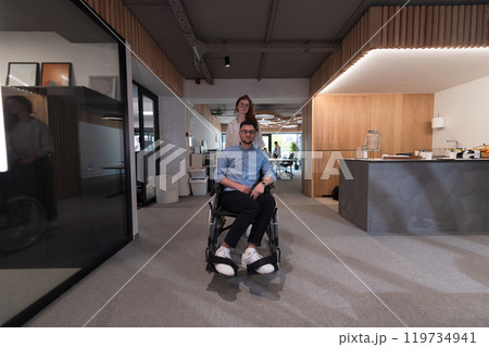 Young business colleagues, collaborative business colleagues, including a person in a wheelchair, walk past a modern glass office corridor, illustrating diversity, teamwork and empowerment in the Young business colleagues, collaborative business colleagues, including a person in a wheelchair, walk past a modern glass office corridor, illustrating diversity, teamwork and empowerment in the 119734941