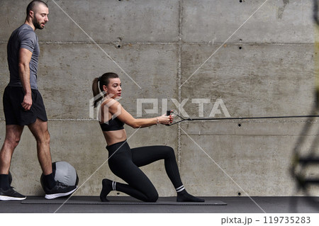 A muscular man assisting a fit woman in a modern gym as they engage in various body exercises and muscle stretches, showcasing their dedication to fitness and benefiting from teamwork and support 119735283
