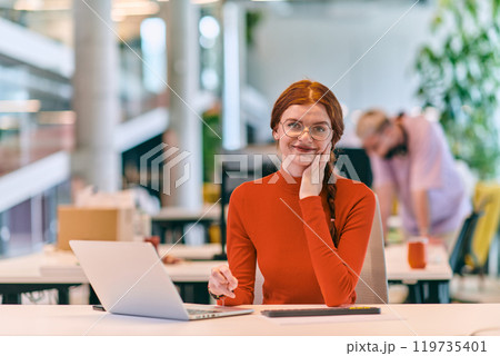 In a modern startup office, a professional businesswoman with orange hair sitting at her laptop, epitomizing innovation and productivity in her contemporary workspace. 119735401