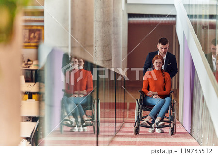 A company director assists his business colleague in a wheelchair, helping her navigate to their startup office, where they work alongside their diverse team of colleagues, emphasizing inclusivity and 119735512