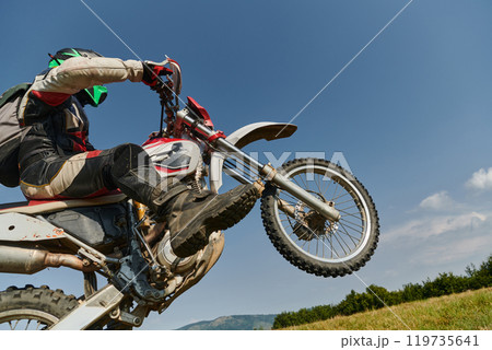 A fearless professional motocross rider skillfully executes an extreme back wheelie maneuver through a perilous forest terrain, showcasing remarkable balance and daring expertise 119735641