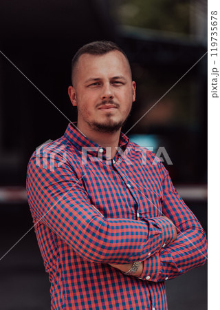 A successful young businessman in a shirt, with crossed arms, poses outdoors, confident expression on his face. 119735678