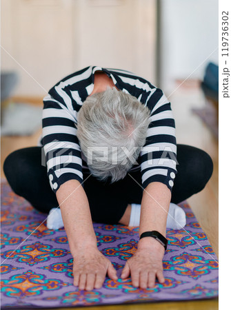 Senior woman performs a back-stretching exercise in a modern, sunlit studio. 119736302
