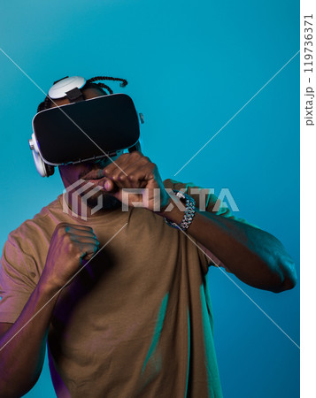 In an avant-garde scene, an African American man engages in cutting-edge virtual reality gaming, utilizing VR glasses to immerse himself in futuristic boxing games, set against a vivid blue background 119736371