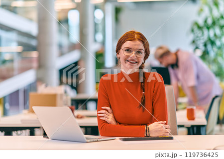 In a modern startup office, a professional businesswoman with orange hair sitting at her laptop, epitomizing innovation and productivity in her contemporary workspace. 119736425