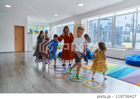 Small nursery school children with female teacher on floor indoors in classroom, doing exercise. Jumping over hula hoop circles track on the floor. 119736427