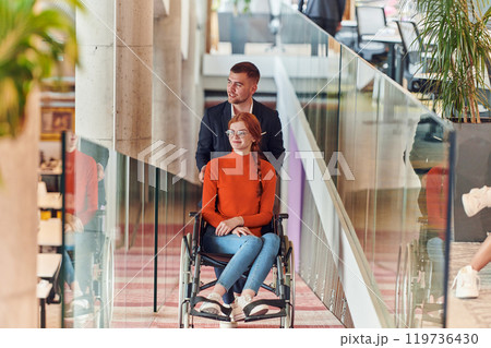 A company director assists his business colleague in a wheelchair, helping her navigate to their startup office, where they work alongside their diverse team of colleagues, emphasizing inclusivity and 119736430