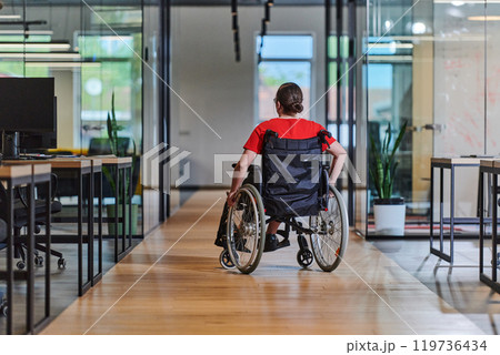 A modern young businesswoman in a wheelchair is surrounded by an inclusive workspace with glass-walled offices, embodying determination and innovation in the business world 119736434