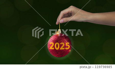 Red Christmas ball with yellow 2025 in woman's hand on green background, New year style 119736985
