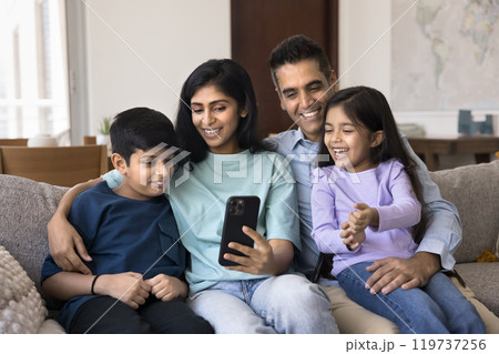 Happy attractive Indian mom and dad using smartphone, hugging kids Happy attractive Indian mom and dad using smartphone, hugging kids 119737256