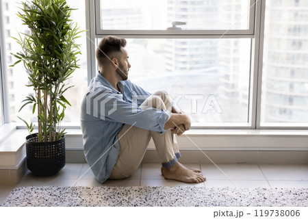 Frustrated lonely Latin man sitting at large window on floor Frustrated lonely Latin man sitting at large window on floor 119738006