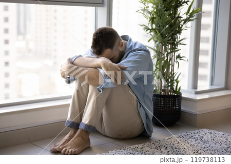 Depressed unhappy young man sitting at window on floor Depressed unhappy young man sitting at window on floor 119738113
