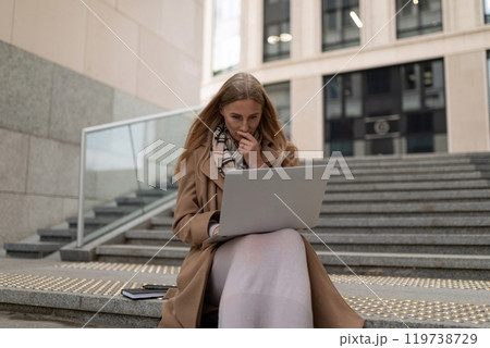 Business woman working intently on her laptop near a modern business center during a sunny day Business woman working intently on her laptop near a modern business center during a sunny day 119738729