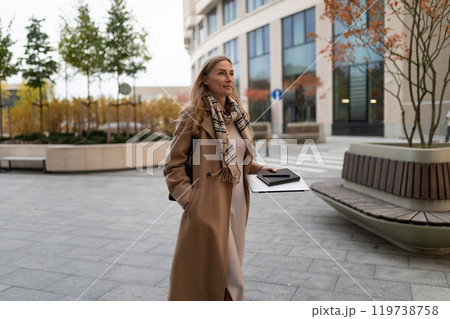 Businesswoman walking confidently near a bustling business center, holding a notebook and looking Businesswoman walking confidently near a bustling business center, holding a notebook and looking 119738758