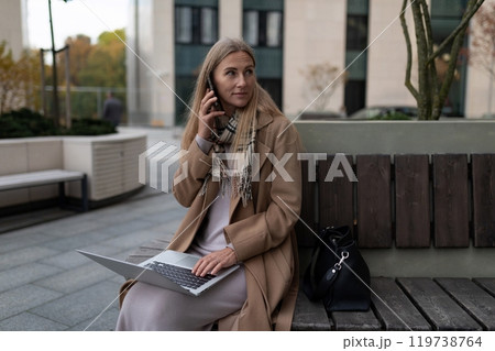 A businesswoman sits on a bench outside a business center, talking on her phone and working on her A businesswoman sits on a bench outside a business center, talking on her phone and working on her 119738764
