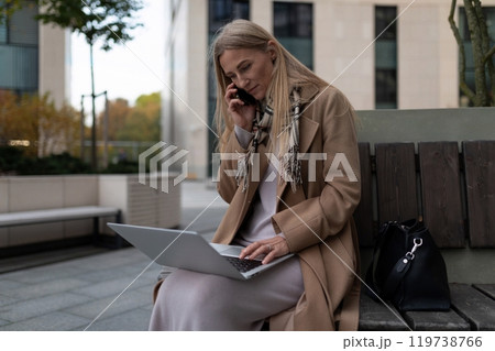 A businesswoman sits on a bench outside a business center, working on her laptop and talking on her 119738766