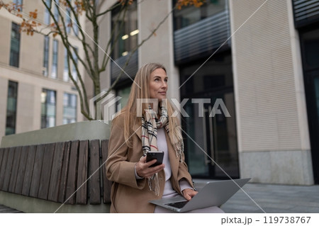 A businesswoman sits on a bench outside a business center, working on her laptop and checking her A businesswoman sits on a bench outside a business center, working on her laptop and checking her 119738767