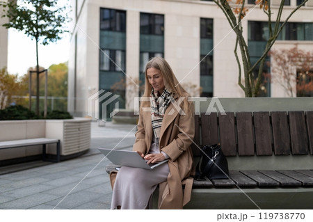 A businesswoman works on her laptop while sitting on a bench outside a business center A businesswoman works on her laptop while sitting on a bench outside a business center 119738770