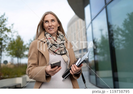 A businesswoman walks near a business center while holding a smartphone and documents 119738795