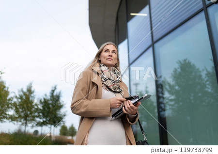 A businesswoman walks by a modern office building while holding a tablet and looking up A businesswoman walks by a modern office building while holding a tablet and looking up 119738799