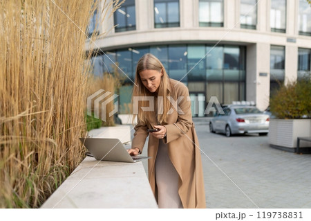 A businesswoman checks her phone while working on her laptop outside a business center A businesswoman checks her phone while working on her laptop outside a business center 119738831