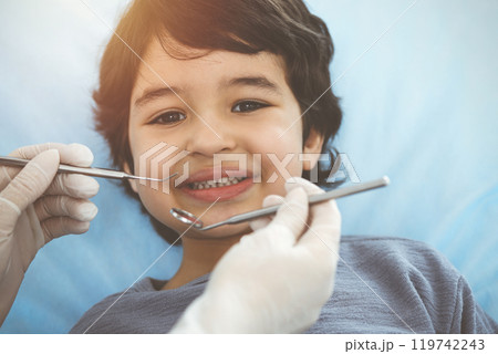 Cute arab boy sitting at dental chair with open mouth during oral checking up with doctor. Visiting sunny dentist office 119742243