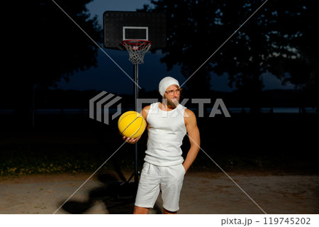Man holding a basketball in a park at dusk, showcasing athleticism and confidence by the court under a darkening sky 119745202