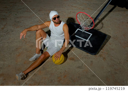 A basketball player sits on the court wearing sunglasses, relaxed after an intense practice session at dusk A basketball player sits on the court wearing sunglasses, relaxed after an intense practice session at dusk 119745219