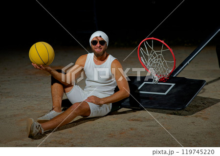A basketball player sits on the court wearing sunglasses, relaxed after an intense practice session at dusk A basketball player sits on the court wearing sunglasses, relaxed after an intense practice session at dusk 119745220