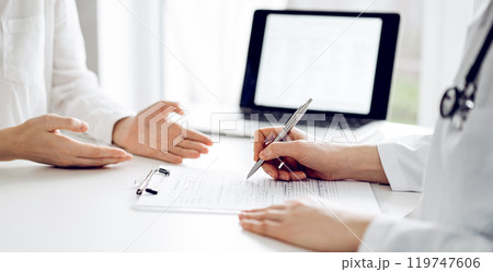 Doctor and patient sitting and discussing something near each other at the white desk in clinic. Female physician is listening filling up a records form. Medicine concept 119747606