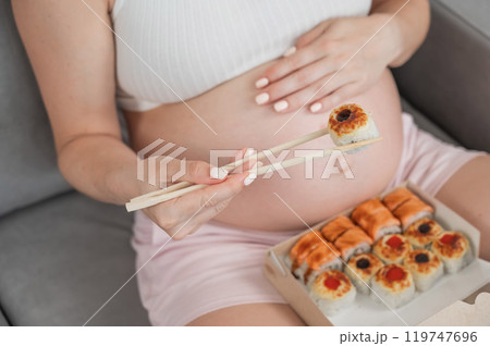 A pregnant woman sits on the sofa and eats rolls from a box. Food delivery. Close up of belly. A pregnant woman sits on the sofa and eats rolls from a box. Food delivery. Close up of belly. 119747696