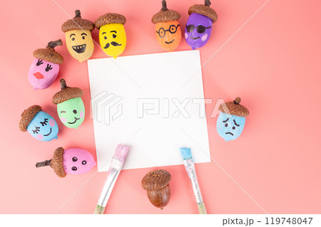 The acorns have brown caps and are painted in colors such as blue, yellow, orange, green, The facial expressions range from happy to sad. The blank white square in the center is empty 119748047