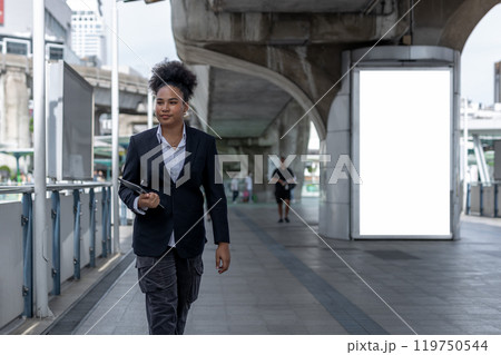 Teenage businesswoman, american african, afro hairstyle Holding a tablet walking on city street rushing to work. Empty white billboard for advertising. Teenage businesswoman, american african, afro hairstyle Holding a tablet walking on city street rushing to work. Empty white billboard for advertising. 119750544