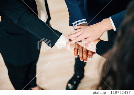 Colleagues from various ethnicities and backgrounds join hands in a modern office, emphasizing teamwork, cooperation, and community. An above view showcases their commitment to equality and diversity. 119752457