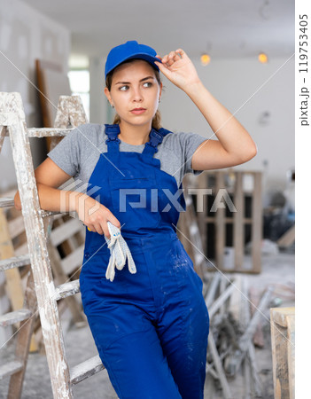 Portrait of contented young female builder in blue overalls during the repair of cottage 119753405