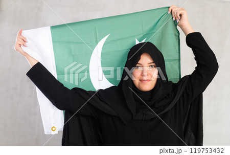 Balanced smiling Muslim woman in traditional black hijab holds flag of Pakistan 119753432