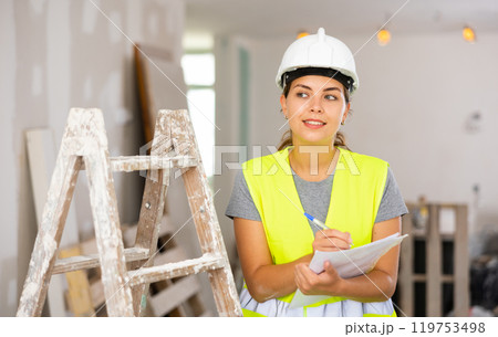 Woman foreman in a protective helmet and yellow vest checks the completed construction work on drawing Woman foreman in a protective helmet and yellow vest checks the completed construction work on drawing 119753498