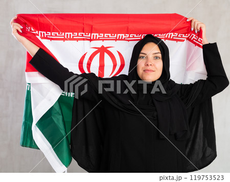 Balanced smiling Muslim woman in traditional black hijab holds flag of Iran 119753523