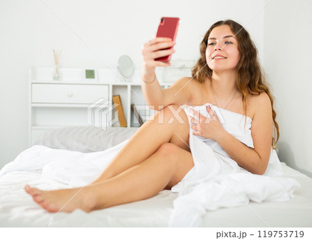 Naked girl covered with sheet taking selfie on bed Naked girl covered with sheet taking selfie on bed 119753719