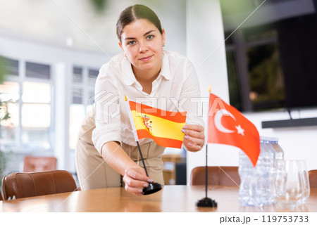 Young female assistant putting up turkish and spanish flags on table for international negotiations Young female assistant putting up turkish and spanish flags on table for international negotiations 119753733