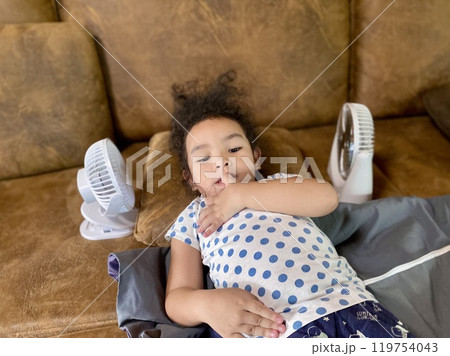 Little Asian girl playing with electric fan on sofa at home 119754043