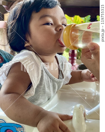 Cute Asian baby girl drinking water from a bottle in the kitchen at home Cute Asian baby girl drinking water from a bottle in the kitchen at home 119755315