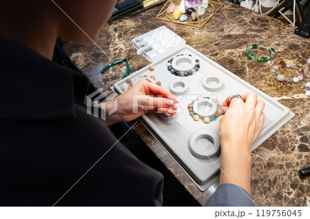 Top view artistic female hands creating bracelet from stones. POV shot of woman working with craft. Accessories on workshop table close up Top view artistic female hands creating bracelet from stones. POV shot of woman working with craft. Accessories on workshop table close up 119756045