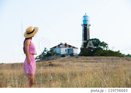 young woman standing in field, nature in background and lighthouse, hipster style. happy girl in straw hat looking at lighthouse 119756083