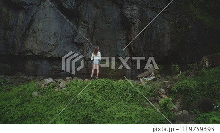 Young woman stands alone, gazing at a massive basalt cliff in a lush tropical forest, lost in thought during an adventurous hike through french polynesia's moorea island 119759395