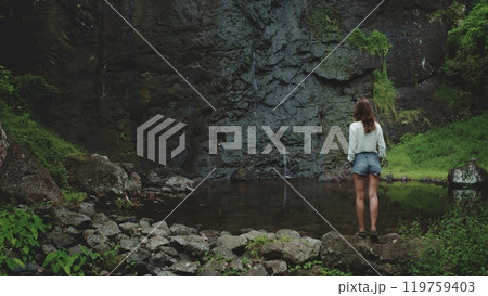 A lone woman stands on a rocky shore, admiring a cascading waterfall in the lush tropical landscape of French Polynesia. The water flows into a tranquil pool surrounded by verdant vegetation. 119759403