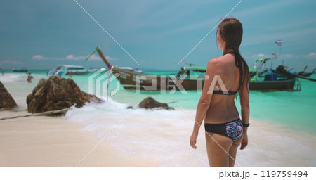 Young woman is enjoying a sunny day on a tropical beach, looking at the horizon where traditional thai longtail boats are moored in the turquoise water. 4K slow motion video 119759494