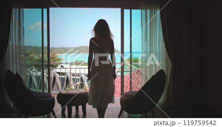 Woman in a flowing white dress walks towards a hotel balcony overlooking the turquoise ocean in southeast asia, enjoying the peaceful and stunning view Woman in a flowing white dress walks towards a hotel balcony overlooking the turquoise ocean in southeast asia, enjoying the peaceful and stunning view 119759542